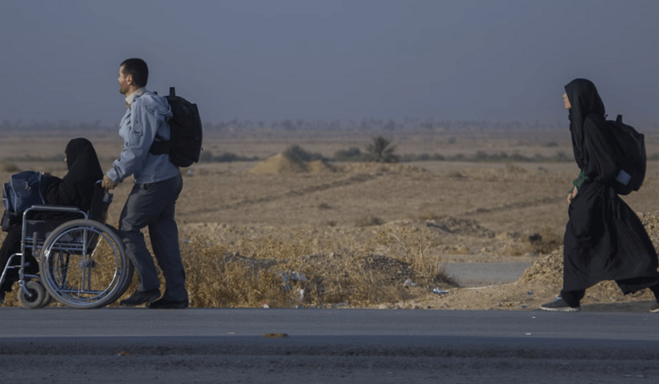 Image shows three people on a roadside with wide, empty expanse of land behind them, one pushing another in a wheelchair.